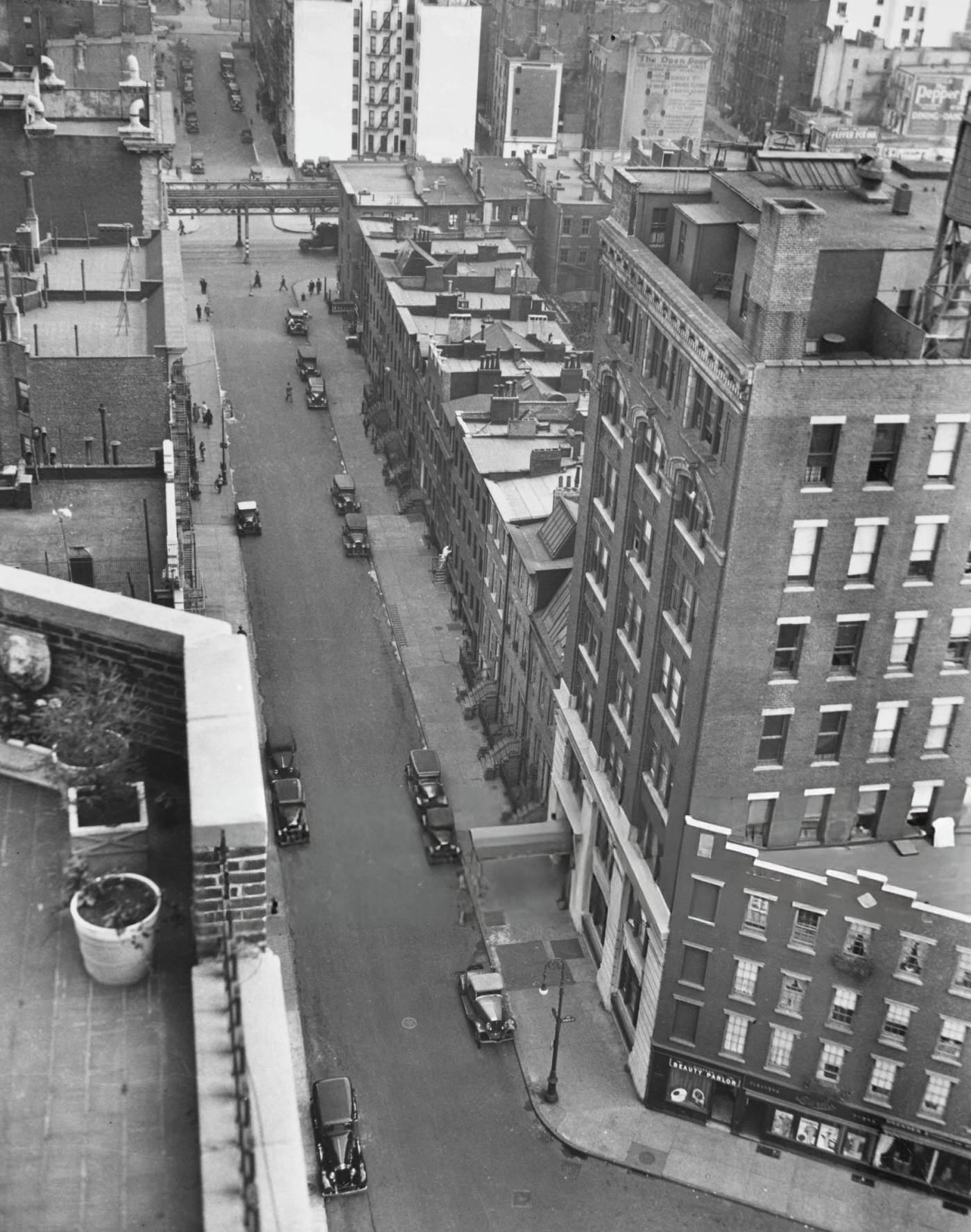 High Angle View Of A Road Between Two Buildings, With Cars Parked Along The Sidewalk, Taken From A Roof Terrace With Potted Plants In Greenwich Village, 1936.