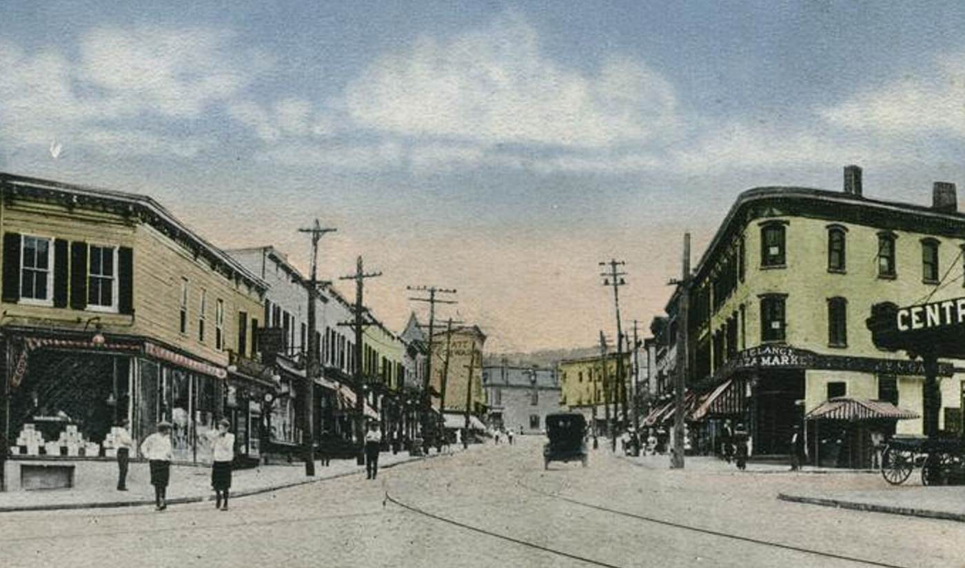 Tompkinsville, with a view of the street with people, markets, an old car, trolley tracks, and shops with awnings, including Plaza Market on the corner.