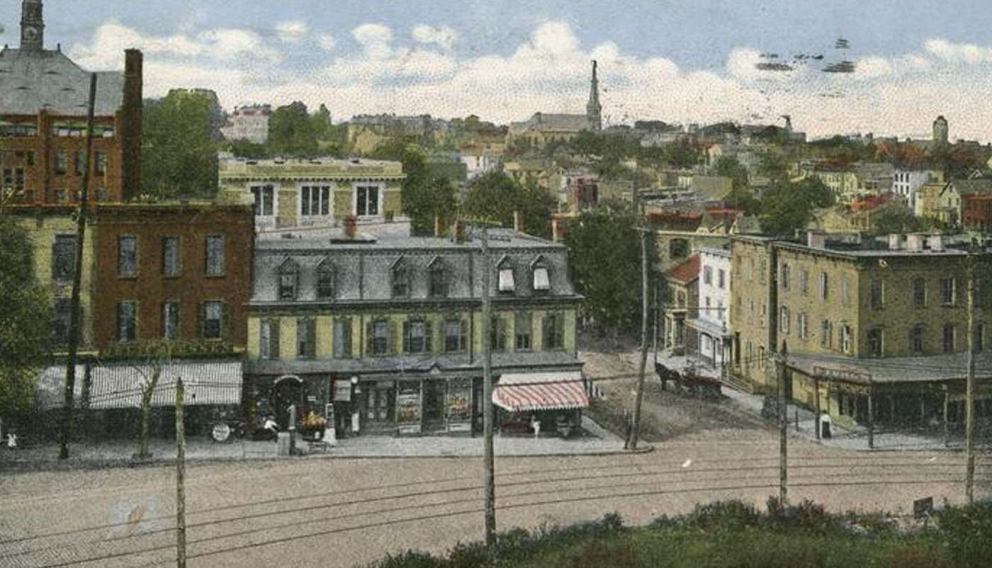 Shopping area in Tompkinsville, with trolley tracks on a brick street, a horse and wagon, and a striped awning over the corner store.
