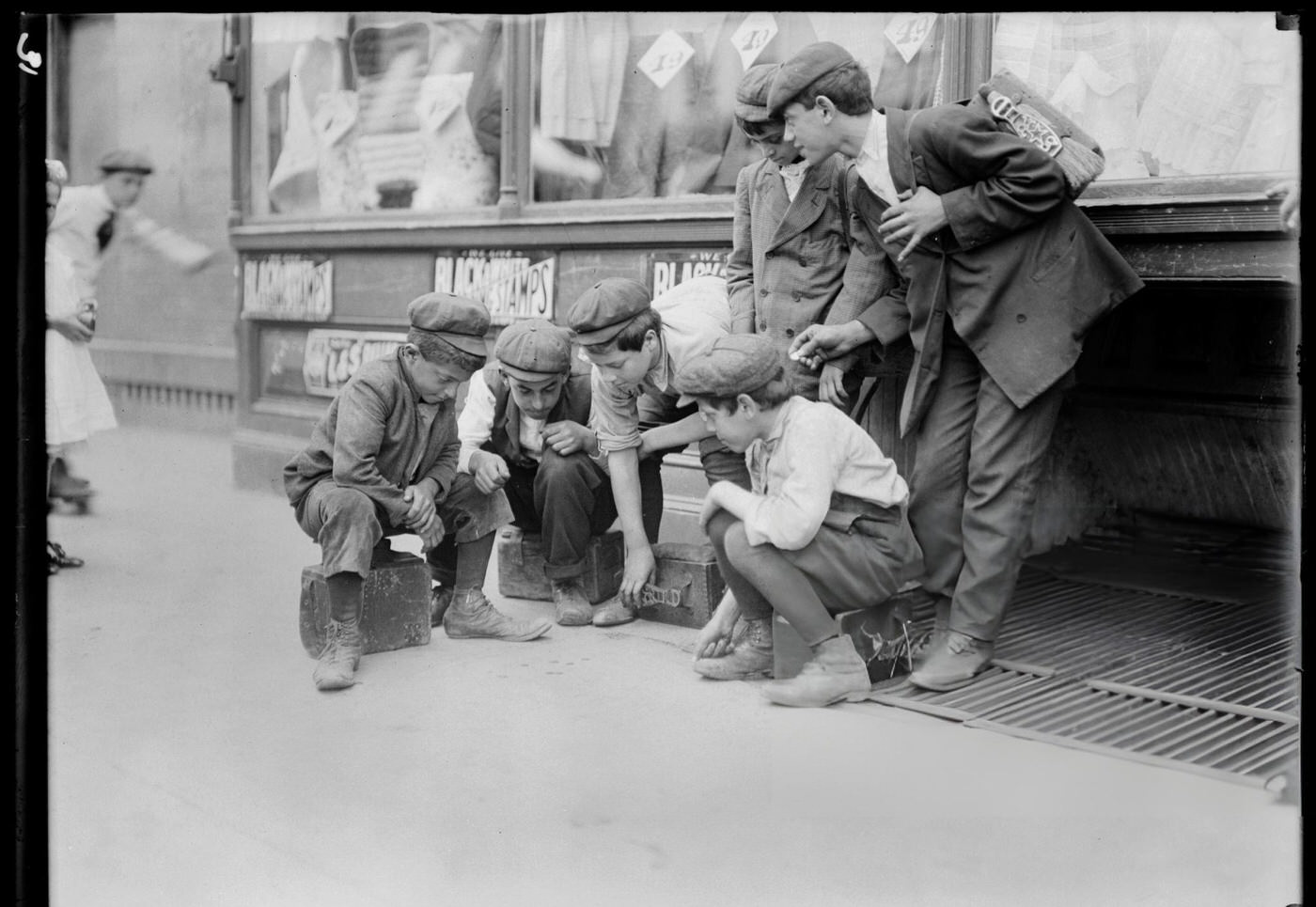 Group Of Newsboys And Shoeshine Boys Playing Craps On A Sidewalk In Front Of A Store, New York, 1910S.