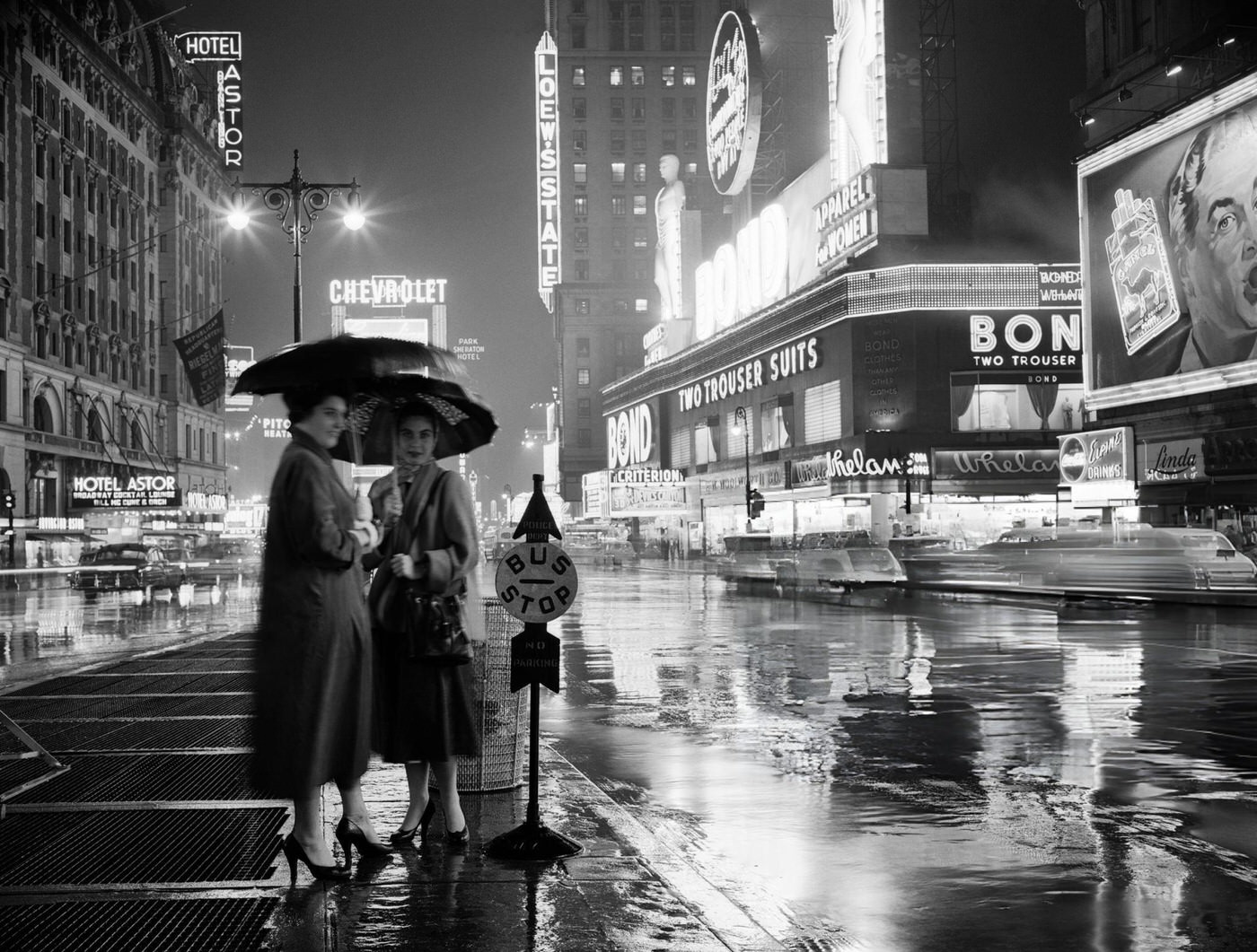 Two Silhouetted Women At A Bus Stop Under Umbrellas On A Rainy Night In Times Square, 1950S.