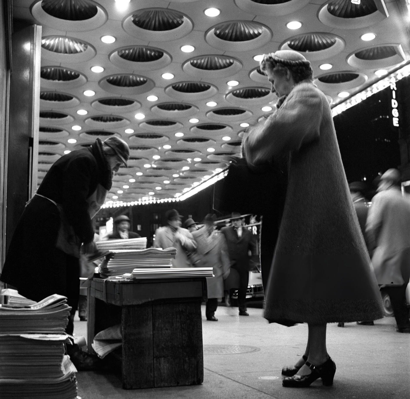 Newspaper Vendor Under The Marquee At The Paramount Theatre In Times Square, With A Customer Reaching Into Her Purse, 1954.