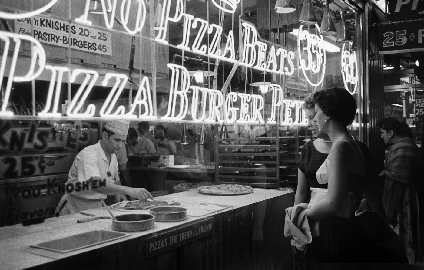 Night Time View Of Two Visitors Watching Pizza Being Prepared By A Chef In The Neon Sign Lit Window Of A Pizzeria And Burger Restaurant In Times Square, 1957.