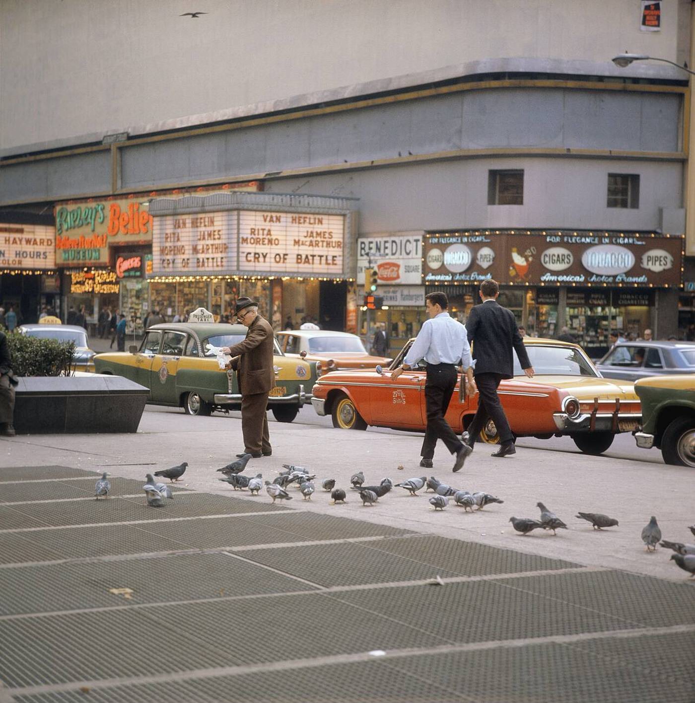 Street Scene With Cars And Taxis Driving On Broadway In Times Square, Showing Movie Theatres And An Elderly Man Feeding Pigeons, 1964.