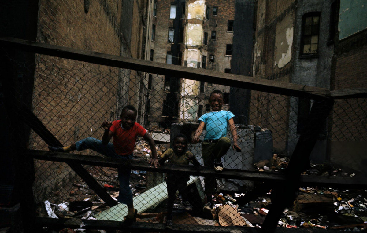 Children Laughing Behind A Fence Near A Vacant Lot And Abandoned Buildings, Harlem, 1970.